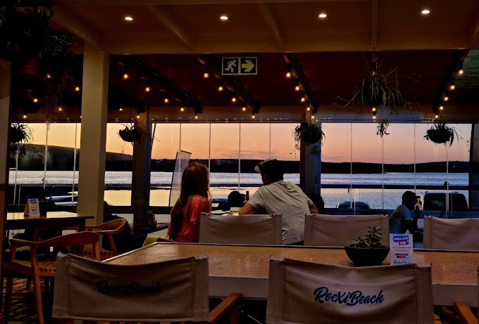 A couplle sits at sunset inside Rocxi beach Restaurant in Langebaan, enjoying the view of the lagoon through glass panels and fairy lights on the ceiling.