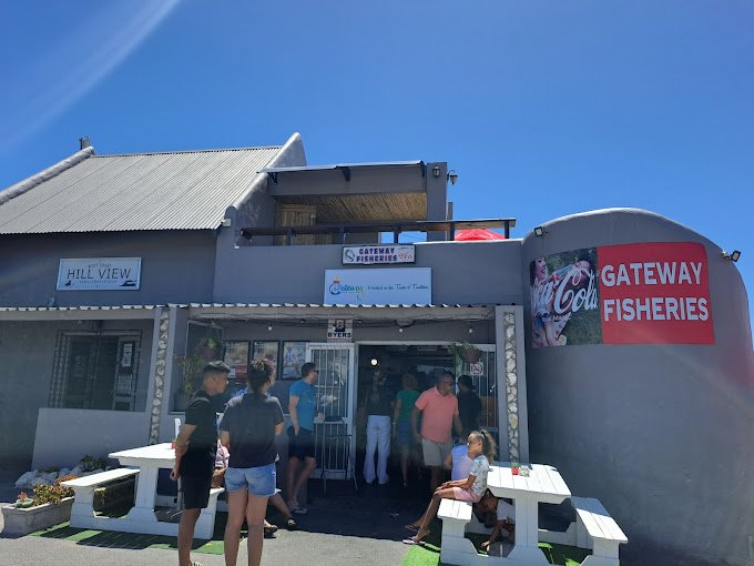 People standing outside and sitting on white benches outside a grey building of Gateway Fisheries in Langebaan. With big sign outside attached to wall, all waiting for fish and chips takeaways.
