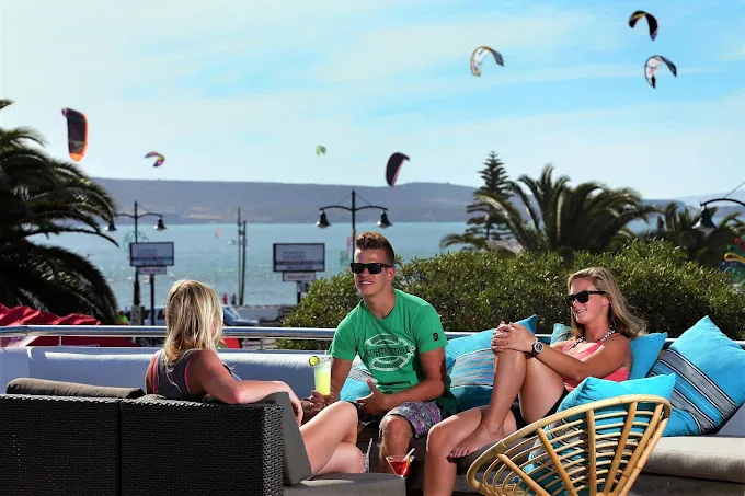 Three young peole sit outside on blue cushions, chatting with the view of the Langebaan lagoon and kite surfers in the background at Breeze Restaurant in Langebaan.