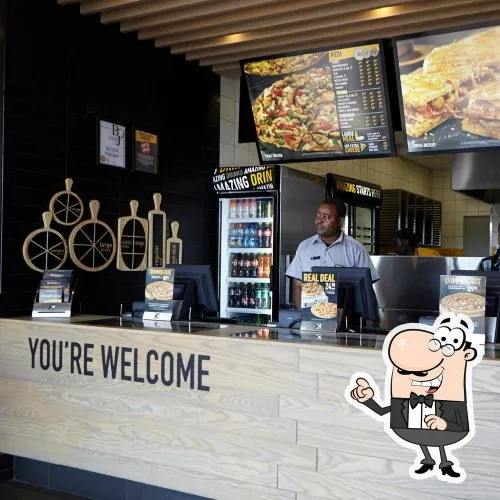 A shop assistant behind a wooden counter at Debonairs Pizza inn Langebaan with digital display boards of the menu attached to the ceiling and fridge with beverages.