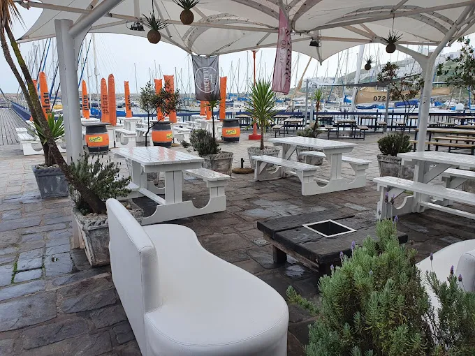 White seating, white tables, white shade covering on slate floors at Dockside Langebaan with a view towards the Marina with lots of boats.