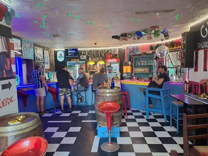 Men standing and sitting at the bar counter at FRIENDS Bar in Langebaan. The bar counte is blue, black and white check floor, wine barrels as tables and colouful chairs all around.