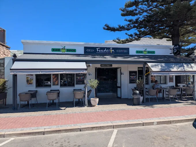 The entrance to the white building of Fooderia in Langebaan with shade covers over tables and chairs outside and signage on the walls.