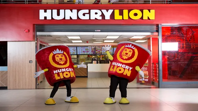 Two mascots in front of Hungry Lion Takeaway in Langebaan. Two mascots dressed like chicken buckets. Exterior is red windows and walls with signage in white and yellow with a view inside towards servinng area.