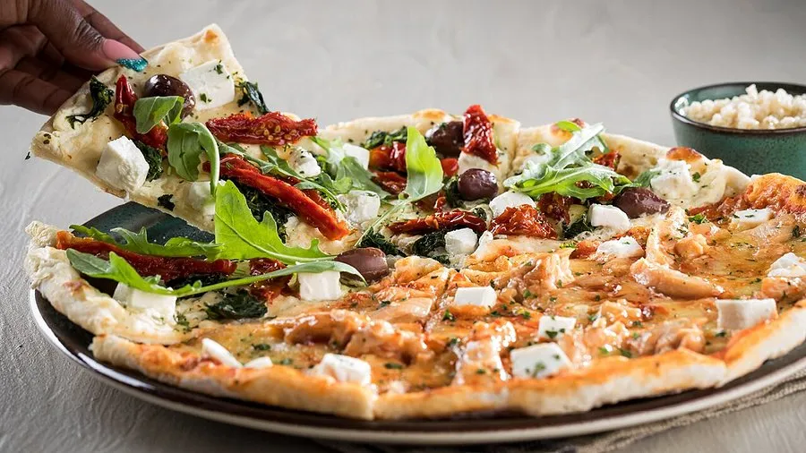 A slice of pizza is being removed from a pizza on a plate, that is coverered with cheese, feta, basil,olives and sunried tomatoes. This pizza is a takeaway from Panarottis in Langebaan.