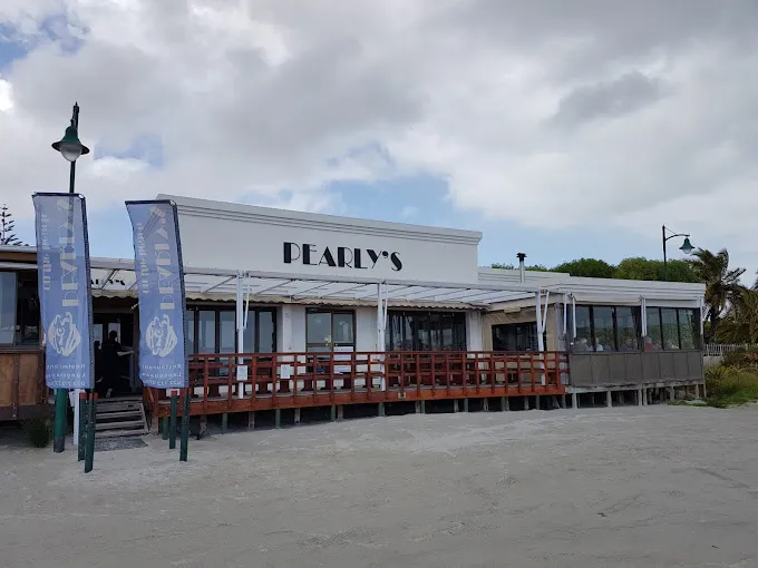 The outside of Pearlys in Langebaan, with beach in front, wooden decck, white building, banners at the entrance and black signage on the walls.