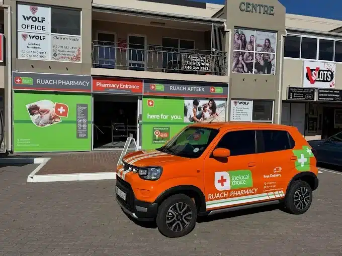 A Orange branded car in front of Rauch Pharmacy in Langebaan with green shopfront windows and signage on bilding.