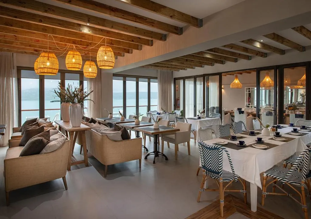 The interior of the Shark Bay Hotel Restaurant. View onto the lagoon with light cream shades of upholstered furniture, chairs and tables with wicked lights, creating a comtempory interior.