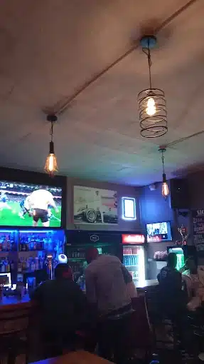 People around a bar counter, at night, watching rugby on big TV screen above fridges with beverages inside, at Sicasa Bar in Langebaan. This is a Sports Bar.