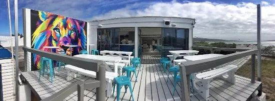 The white wooden deck of Wunderbar Langebaan with colourful wall murual of a lion and lots of white tables, benches, and bright colour chairs. Great view of the lagoon in a distance.