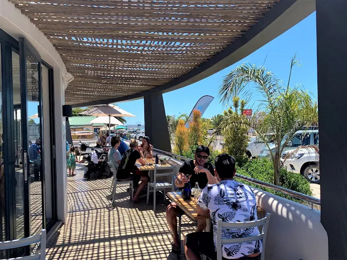 People sitting under wooden ceiling on the outside deck of the Breeze Restaurant in Langebaan with wooden floors, greenery and view towards lagoon along Bree Street.