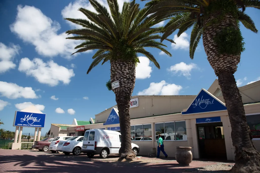 The exterior of The Watergat in Langebaan with aprking and two palm trees in front of building with blue signage.