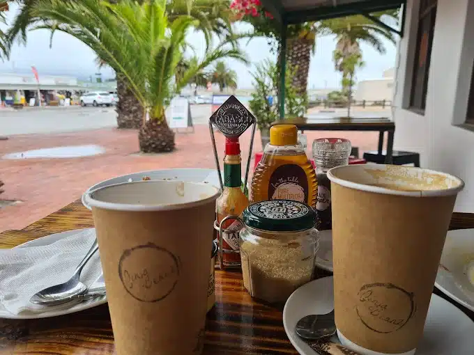 Coffees on the outside table with condiments needed at Ginja Beanz Coffee Cafe in Langebaan. Close to the main beach with palm trees in a distance towards the Langebaan lagoon.