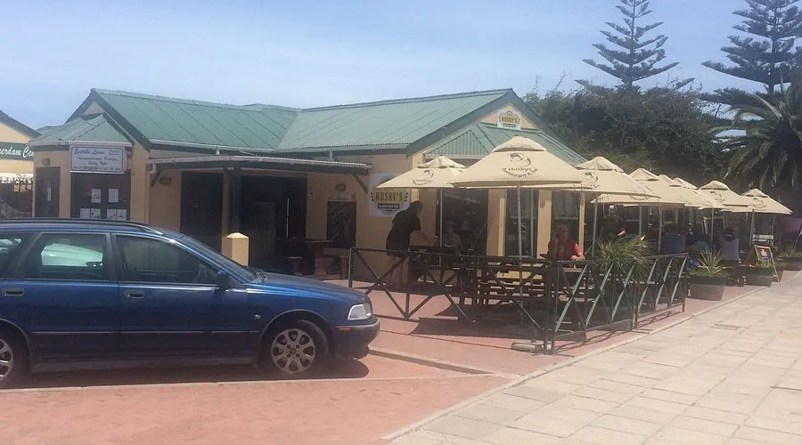The street view towards Husky's Resaurant Langebaan with car parked infront of cream building, green roof and doors and windows and seating outside under umbrellas.