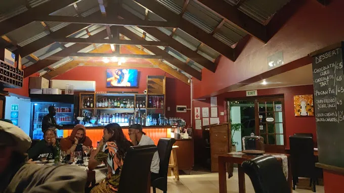Interior of the Queens Cottage Restaurant in Langebaan showing wooden bulkheads, people sitting around table on black chairs and serving counter with red background.