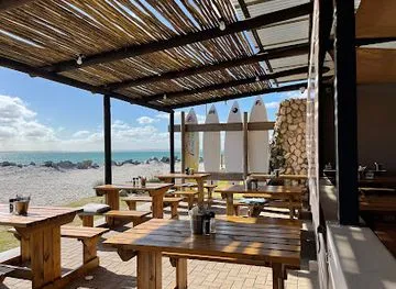 The outside deck of Kokomos langebaan restaurant with wooden tables and chairs, close to the beach, wooden canopy and white surfboards displayed as fenching.
