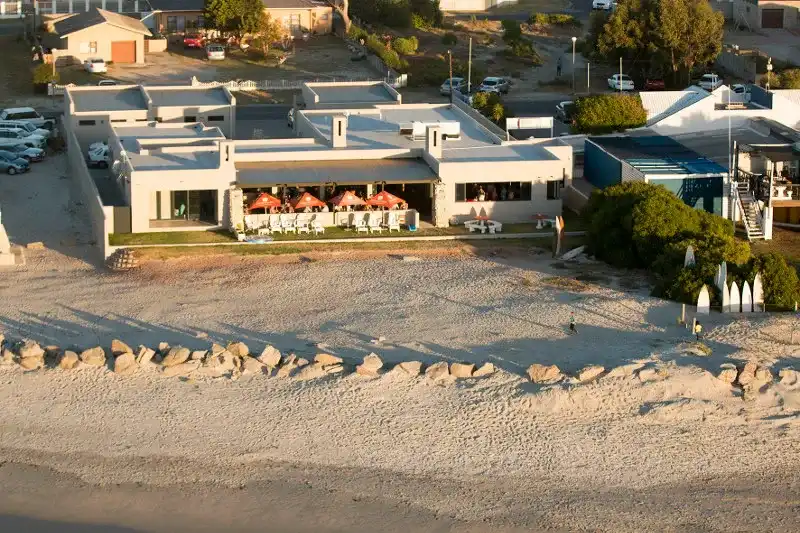 A distance view of the Kokomos Restaurant in langebaan showing white buildinng, umbrellas outside on deck and lots of beach sand on the beach at Langebaan.