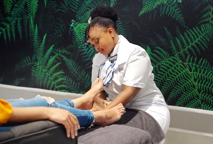 A La Vita Spa assistant does a foot treatment to a customer as they client is sitting a grey chair facing towards wallpaper with green ferns on the wall.