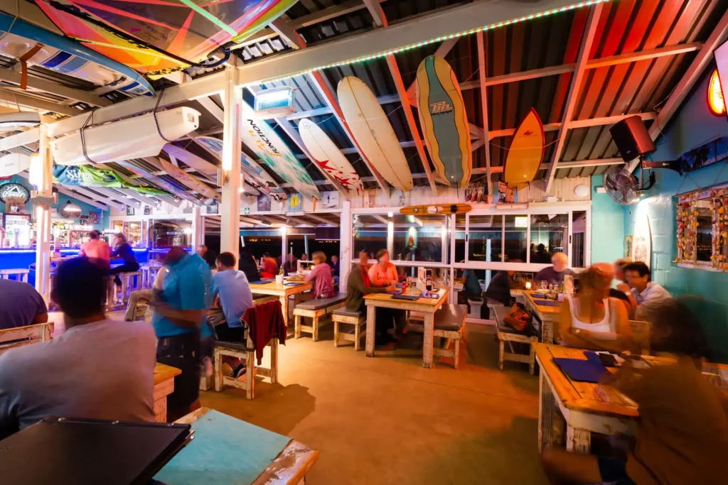 The interior of Marc's Beach Bar at night with warm colours , surfboards attached to the ceiling and peole sitting at tables
