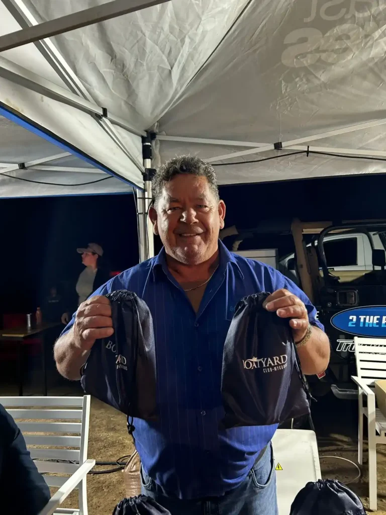 A picture of Nollies, the proud owner of The Boatyard in Langbeaan with two promotional bags in his hands.