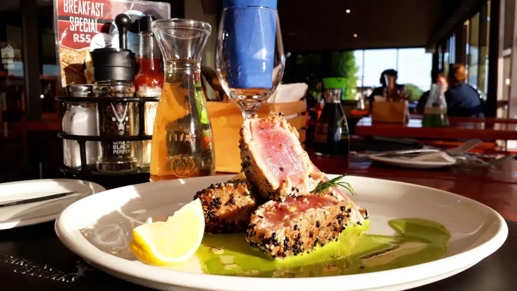 On a white plate you find steak seasoned with lemon and green sauce with glassware on table and people sitting in a distance at Pearly's Langebaan Restaurant on the beach.