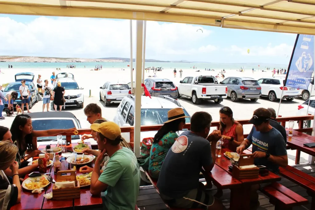 Lots of people sit on outside deck under shade at Pearly's Langebaan restaurant on the beach, enjoying lunch.