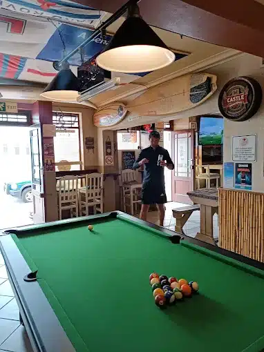 Inside the Sand Bar in Langebaan you have a man standing next to green pooltable and interior of Bar with surfboards, sails and other merchandise on display.