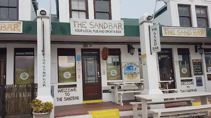The outside of The Sand Bar in the Marra's Building in Langebaan with outdoor seating and lots of signage on walls and windows.