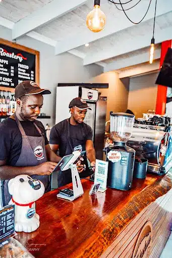 Two staff members behind serving counter preparing coffee reats at Weskus Coffee Roastery in Langebaan with lots of machines on counter and menu on blackboard against the wall.