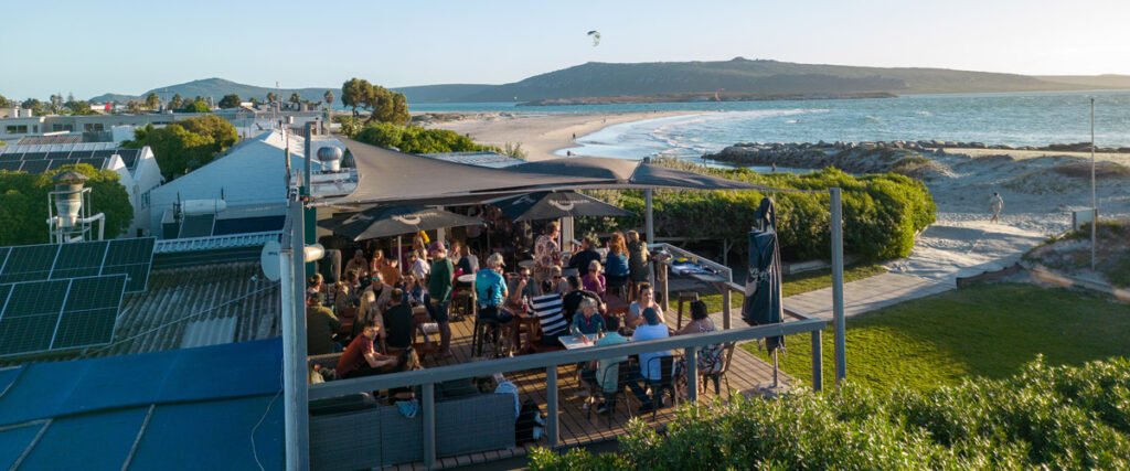 Lots of people on the outside deck at Wunderbar Langebaan with green lawn, sea sand and lagoon in adistance. All creating a great view from terrace.