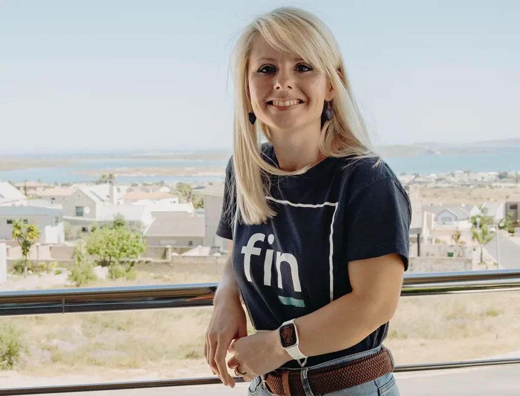 Smiling blonde woman in a black 'fin' T-shirt leaning on a railing with a coastal town and sea in the background.