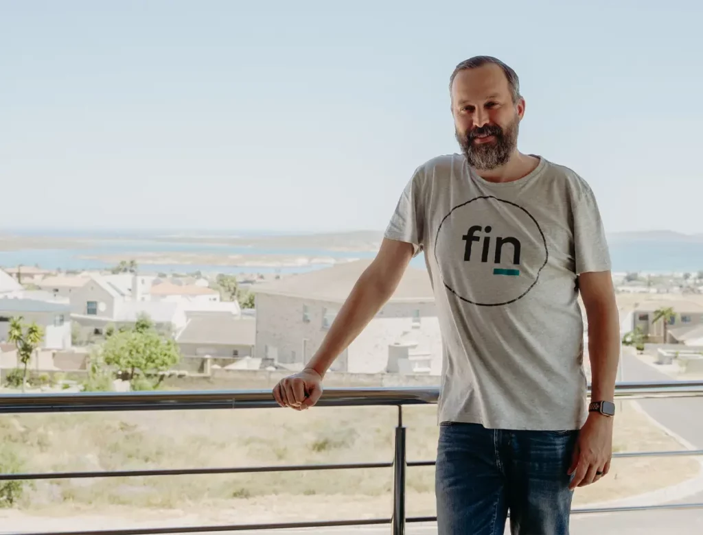 Bearded man in a light gray t-shirt with a circular 'fin' logo stands on a balcony overlooking a coastal town by the sea.